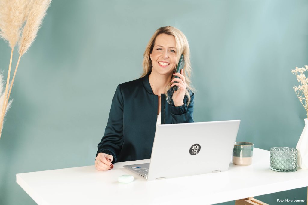 Iris Schulz sitzt telefonierend vor einem Laptop und Lacht dabei in die Kamera